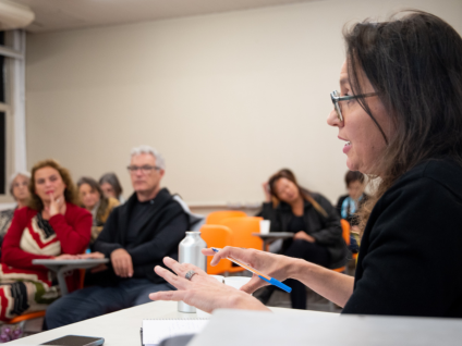 Foto de uma mulher em uma sala de aula fazendo uma apresentação para um grupo de pessoas. Ela é branca, tem cabelos lisos, longos e castanhos, usa óculos e veste uma blusa preta. Ela está sentada, olhando para frente e falando, com uma caneta em uma das mãos. Ao fundo, desfocados, oito pessoas estão sentadas em cadeiras laranjas a observam. As paredes são beges.