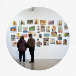 um homem e uma mulher, de costas, observam uma conjunto de pinturas redondas, quadradas e retangulares