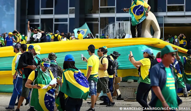 Foto de várias pessoas vestidas de verde e amarelo, com bandeiras do Brasil e uma faixa grande com listras verdes e amarelas, em volta e em cima da estátua da Justiça. Alguns deles estão com o celular em mãos. Ao fundo, um prédio com vidros quebrados e manchados.