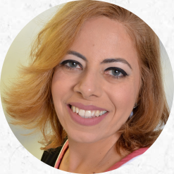 Foto de uma mulher branca de cabelos loiros e lisos na altura do pescoço. Ela tem olhos verdes, está maquiada e sorri, olhando diretamente para a câmera.
