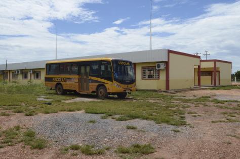 Foto da escola localizada no quilombo Mata Cavalo. O prédio é comprido e possui paredes amarelas, com listras vermelhas e muitas janelas. Na frente da construção, há um ônibus escolar amarelo. O chão é de terra e há nele muitas pedras, além de alguns pontos com grama.