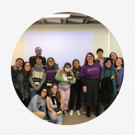 um grupo de estudantes posa reunido para uma foto, a maioria de pé, enquanto duas jovens estão agachadas.. Ao fundo, uma parede branca em que é projetada uma luz azul, retangular.
