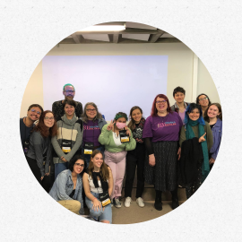 um grupo de estudantes posa reunido para uma foto, a maioria de pé, enquanto duas jovens estão agachadas.. Ao fundo, uma parede branca em que é projetada uma luz azul, retangular. 