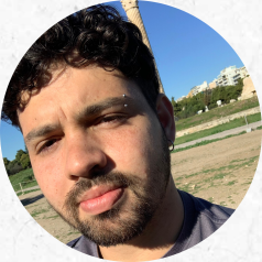 Foto de um homem branco de cabelos cacheados, curtos e escuros e com barba e bigode. Ele utiliza um piercing na sobrancelha e brinco de argola na orelha. Atrás dele há uma paisagem com grama verde, terra, céu azul e alguns prédios.
