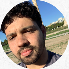 Foto de um homem branco de cabelos cacheados, curtos e escuros e com barba e bigode. Ele utiliza um piercing na sobrancelha e brinco de argola na orelha. Atrás dele há uma paisagem com grama verde, terra, céu azul e alguns prédios. 