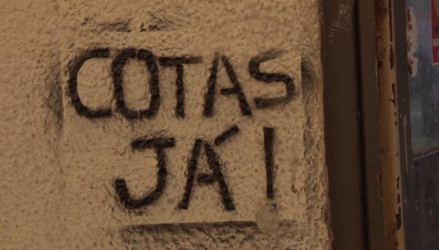 Foto de uma parede bege texturizada com a frase “Cotas já!” escrita em stencil com tinta preta. Do lado direito, o batente de uma porta marrom. A imagem aparenta ter sido captada à noite.