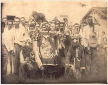 Fotografia antiga em sépia de brincantes da Folia de Reis segurando estandartes, bandeiras e instrumentos musicais. Nela há um grupo de pessoas, majoritariamente homens posando em um terreno gramado. A maioria utiliza camisa social branca e calça escura e se encontra em pé. Uma mulher que está centralizada na imagem segurando uma bandeira enquanto outras duas pessoas estão agachadas ao seu lado utilizando máscaras e adornos.