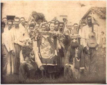 Foto em branco e preto de brincantes da Folia de Reis segurando estandartes, galhardetes, uma espécie de bandeira, e instrumentos musicais. Há uma mulher ao centro, atrás da bandeira, cercada por homens. Agachados ladeando a bandeira, há duas pessoas com máscaras e roupas com adornos.