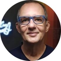 Foto de homem branco de cabelos curtos grisalhos. Ele sorri e usa óculos de armação retangular azul e camiseta preta, o fundo é escuro.