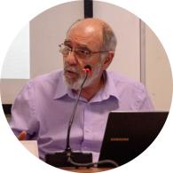 Foto de homem branco sentado, falando em microfone. Ele é calvo, tem barba e cabelos curtos e grisalhos,  olha para a esquerda da imagem, usa óculos e veste uma camisa lilás. À sua frente há um notebook e, no  fundo, uma parede clara e uma tela com projeção. 