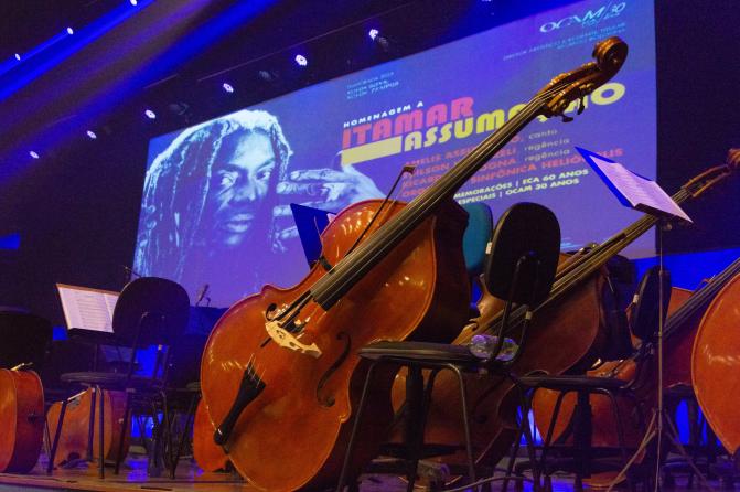 Foto tirada do palco, com foco em contrabaixos dispostos à frente de cadeiras vazias. Ao fundo, há uma grande projeção que ocupa toda a parede, com o rosto de um homem negro com dreads longos e uma das mãos levantada próxima ao rosto. A imagem é acompanhada por texto colorido com nomes, títulos e informações sobre o concerto nas cores vermelha e amarela, principalmente. A iluminação é predominantemente azul, com refletores voltados para a frente do palco.
