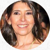 Foto de mulher branca sorrindo. Ela tem cabelos compridos, castanhos e lisos, usa um colar amarelo e maquiagem.