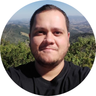 Foto de homem branco de olhos castanhos, cabelos curtos, lisos e castanhos e barba aparada esboçando sorriso. Ele veste camiseta preta. Ao fundo, há uma paisagem com morros, árvores e o céu azul.