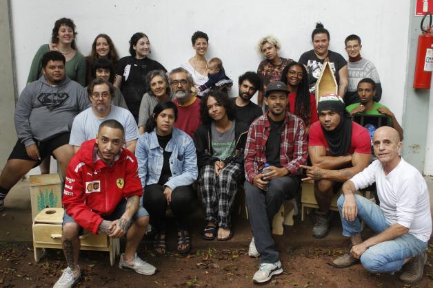  Na foto, cerca de 20 pessoas estão sorrindo, em pé ou sentadas em banquinhos de madeira dispostos em um chão de terra. São pessoas brancas e negras com idades variadas. Ao fundo, uma parede branca e um extintor de incêndio.