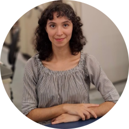 Foto de mulher branca de olhos castanhos, cabelos castanhos e cacheados na altura dos ombros, com franja, sentada diante de uma mesa azul com as mãos sobrepostas. Ela usa uma blusa acinzentada e listrada e há estantes móveis ao fundo.