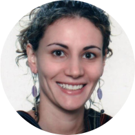 Foto de mulher branca, sorrindo. Ela tem cabelos escuros e ondulados, que estão presos, olhos castanhos, usa brincos pendentes, veste blusa escura. O fundo é branco.