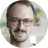Foto de homem branco sorrindo. Ele tem cabelos curtos, lisos e castanhos, bigode e pouca barba. Ele usa óculos de armação redonda marrom rajada e camiseta preta. O fundo tem uma parede cinza e elementos coloridos desfocados.
