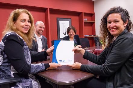 Foto de duas mulheres - ambas brancas, uma de cabelos longos e loiros e outra de cabelos castanhos cacheados - sorrindo enquanto posam segurando um documento com capa azul e branca, com os logotipos "eca" e "USP" em azul. Atrás delas, um homem branco careca e com barba e uma mulher amarela de cabelos castanhos e curtos também sorriem. Todos estão sentados em torno de uma mesa retangular de madeira. Ao fundo, uma parede vermelha com prateleiras embutidas e um quadro.