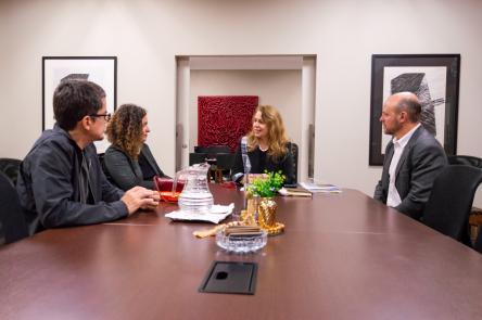 Foto de quatro pessoas - dois homens brancos e duas mulheres brancas - conversando, sentados em torno de uma mesa retangular de madeira. Um dos homens tem cabelo escuro e curto, usa óculos e uma jaqueta preta. A mulher ao lado dele tem cabelos longos, cacheados e castanhos e usa um blazer preto. Voltada para essa, uma mulher com longos cabelos loiros e ondulados usa um blazer preto. O outro homem é calvo, tem barba e veste um blazer cinza. Ao fundo, uma janela com persiana clara e uma parede bege com um quadro abstrato.