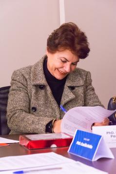 Foto de uma mulher branca, de cabelos curtos, lisos e castanhos, vestindo um blazer cinza e uma blusa preta, assinando um documento. Com uma das mãos ela segura a caneta e com a outra, as folhas de papel. À sua frente, em cima da mesa, estão uma pequena caixa vermelha, uma caneta e outros papéis. Ao fundo, uma parede bege.