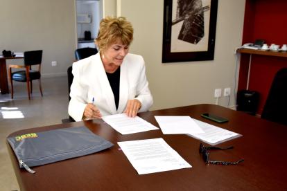 Foto de uma mulher branca, loira e de cabelos curtos, vestindo um blazer branco e uma blusa preta. Ela está sentada em uma cadeira, em frente de uma grande mesa de madeira marrom escura. Ela segura algumas folhas de papel, olhando para elas, e uma caneta. ao fundo, uma parede na cor bege, em que está pendurada uma obra de arte, e cadeiras do escritório.