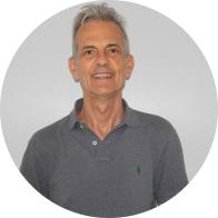 Foto de foto de homem branco com olhos escuros e cabelos curtos e grisalhos sorrindo. Ele veste uma camiseta polo cinza e o fundo da imagem é cinza. 