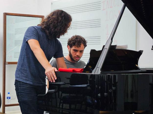Foto de dois homens brancos e um piano de cauda preto. Um deles, de cabelos curtos e cacheados e barba castanhos e camiseta cinza, está sentado à frente do piano e o outro, de cabelos longos, cacheados e castanhos, camiseta azul e calça jeans, está de pé ao lado dele. Em cima do piano há um tecido vermelho. No fundo, paredes brancas com uma lousa e um espelho pendurados.
