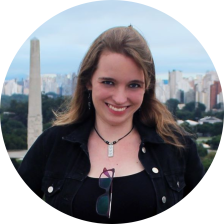 Foto de uma mulher branca sorrindo. Ela tem cabelos castanhos, lisos e compridos, olhos escuros e lábios rosados. Ela usa jaqueta e camiseta pretas, com um óculos pendurado na roupa e um colar. Ao fundo, um obelisco, árvores, prédios e um céu nublado.