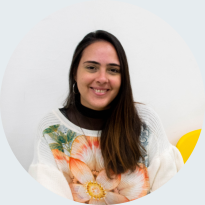 Foto de uma mulher branca sorrindo, de cabelos castanhos, lisos e longos. Ela usa uma blusa branca com estampa florida em tons de laranja, verde e amarelo. O fundo é branco com um detalhe em amarelo.