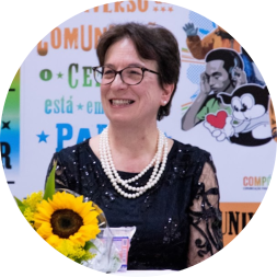 Foto de mulher branca sorrindo. Ela tem cabelos castanhos, lisos e curtos, veste uma blusa preta com brilho e mangas translúcidas, usa óculos de armação preta, brincos e colares de pérolas. À sua frente, um girassol. Ao fundo, um painel com escritos em laranja, verde e azul.
