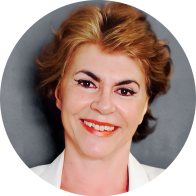 Foto de uma mulher branca sorrindo. Ela tem cabelos avermelhados, lisos e curtos, olhos castanhos, veste blusa branca e usa batom vermelho.  Atrás dela, um fundo cinza.