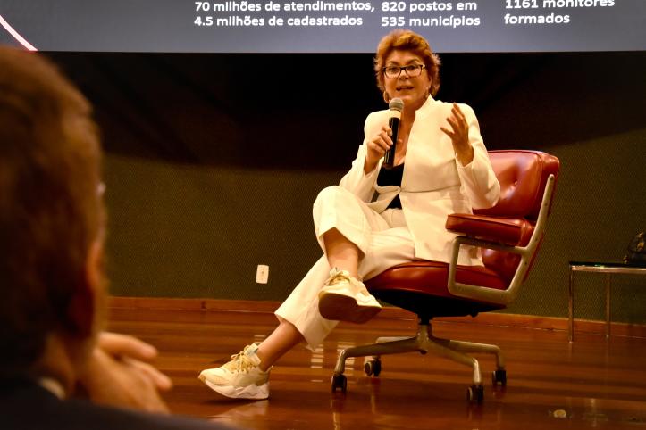Foto de uma mulher branca sentada em uma cadeira vermelha falando ao microfone com as pernas cruzadas. Ela tem cabelos curtos, lisos e loiros escuros, está de tênis e usa calças e paletó brancos, blusa preta e óculos. Atrás dela, uma parede escura e parte de uma projeção com números. O piso é de madeira. No primeiro plano da foto, que está desfocado, há uma cabeça de costas para a câmera e de frente para a mulher. 