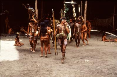 Frame de um filme. Na imagem, um grupo com alguns indígenas adultos com pinturas corporais. Os homens possuem cabelos lisos, pretos e curtos até a altura das sobrancelhas e as mulheres têm os cabelos longos, lisos e pretos até a cintura. À frente, um homem com um arco de caça. O chão é de terra batida e no fundo há estruturas de madeira como colunas.