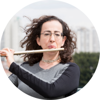 Foto de mulher branca tocando flauta transversal. Ela tem cabelos ondulados, longos e escuros, que estão esvoaçantes, e usa óculos com lentes retangulares. Ela veste uma blusa cinza e preta e usa um colar prateado. Ao fundo, prédios e vegetação desfocada.