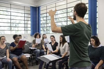 Um jovem branco, de cabelos curtos e castanhos e barba da mesma cor, está de pé, com os braços ao alto. à sua frente uma partitura e mais ao fundo, um grupo de jovens estão sentados, com folhas nas mãos, cantando. Ao fundo, as janelas da sala de aula.