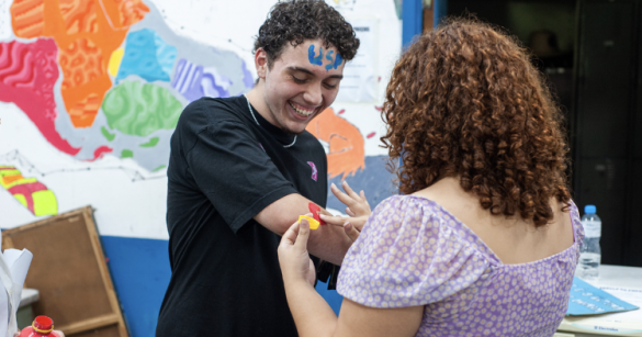 Foto de um homem branco sorrindo enquanto uma mulher branca escreve algo no braço dele com tinta vermelha. O homem tem cabelos pretos, cacheados e curtos, usa uma camiseta preta e tem a palavra USP escrita na testa com tinta azul. A mulher está de costas para a câmera, tem cabelos ruivos e cacheados e usa uma blusa lilás com bolinhas brancas. No fundo, uma parede colorida.