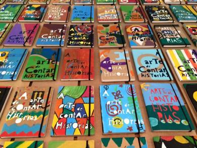 Foto de livros cartoneros. Há cerca de 42 livros enfileirados sobre uma superfície marrom. Todos têm o mesmo título e as capas são coloridas e diferentes.