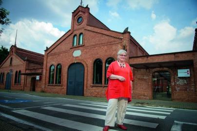 Foto de Maria Dora Mourão, uma mulher de cabelos brancos e curtos. Usa óculos de haste vermelho escuro, colar e tênis da mesma cor, calça bege e blusa vermelha. Na foto, está de pé, na frente do edifício-sede da Cinemateca Brasileira. O prédio antigo é feito de tijolos aparentes, em cor de barro, com portas e janelas em forma de arco.