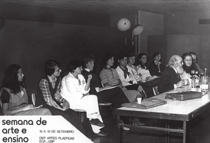 Fotografia em preto e branco. Na porção esquerda da imagem, tem início uma fileira de pessoas que assistem ao evento e se estende até o plano de fundo e a porção direita da imagem. À frente dessa fileira, sentados de frente a uma mesa grande, é possível ver Paulo Freire, um senhor idoso, calvo, de pele branca, cabelos grisalhos e barba branca espessa. Ele usa um óculos quadrado, terno e gravata e fala diante de um microfone. Ao seu lado está a professora Ana Mae Barbosa, uma mulher branca de cabelos escuros e espessos que usa uma blusa de cor clara, além de um homem não identificado, já que seu rosto está quase totalmente coberto pelo rosto de Ana Mae. Na parte inferior esquerda da imagem, a legenda Semana de Arte e Ensino. 15 a 19 de setembro. Departamento de Artes Plásticas. ECA USP. 