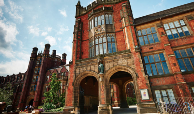 Foto da fachada de um dos prédios da Universidade de Newcastle, no Reino Unido. O prédio é antigo e tem grandes janelas de vidro azul. Ele é feito de tijolod avermelhados. Na entrada, há dois portais. À esquerda da imagem há uma árvore pequena e à direita há um bicicletário. Ao fundo da foto, no canto superior esquerdo, vê-se o céu, em um tom azul claro com poucas nuvens.