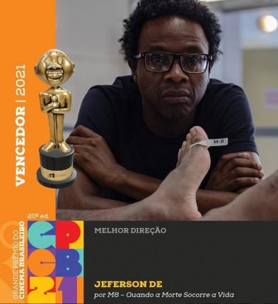 Arte oficial da 20ª edição do Grande Prêmio do Cinema Brasileiro mostra fotografia do diretor Jefferson De, um homem negro, de cabelos crespos pretos e curtos e óculos de armação retangular na cor preta. Ele usa uma camiseta preta e olha para a câmera de braços cruzados. Na sua frente aparecem os pés de um homem negro, identificado pela etiqueta M-8 presa em um dos dedões. Na parte inferior da imagem aparecem o logo do Prêmio e os dizeres em letras brancas e amarelas sobre um retângulo preto: Melhor Direção: Jefferson De, por M8 - Quando a Morte Socorre a Vida. Na parte esquerda da imagem, sobre um fundo retângulo laranja em letras brancas, aparecem os dizeres Vencedor 2021 ao lado de uma reprodução do Troféu Grande Otelo.