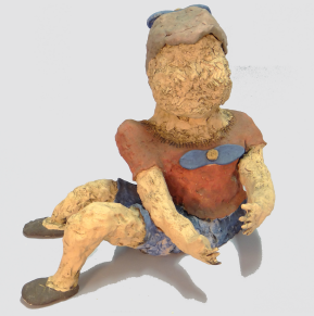 Foto de um boneca feita de argila. A figura, que não tem traços faciais, está sentada e com os braços estendidos para o lado esquerdo. O cabelo é cinza e é enfeitado com um laço azul na parte de trás. A boneca veste uma blusa vermelha com laço azul, um short azul e sapatos cinzas.