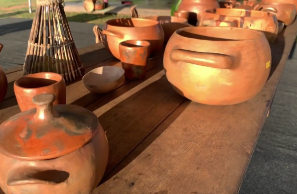 Vários objetos de cerâmica feitos com barro, como panelas, copos e tigelas, estão expostos em uma mesa de madeira. Algumas das peças têm manchas escuras.