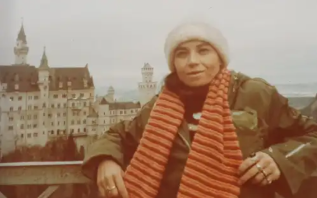 Foto de uma jovem sorrindo para a câmera. Ela é branca e tem seus cabelos escuros cobertos por uma touca branca. A moça veste um cachecol com listas vermelhas e laranjas e um casaco verde. Ao fundo, uma construção antiga, que lembra um castelo.