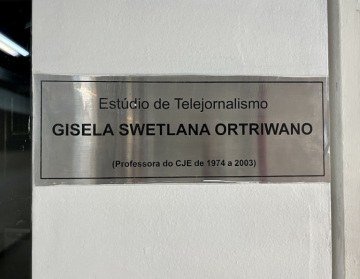 Foto de uma placa fixada em uma parede branca. A superfície da placa é prata e um pouco reflexiva. Em letras pretas, está escrito: Estúdio de Telejornalismo Gisela Swetlana Ortriwano (Professora do CJE de 1974 a 2003).