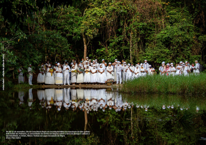 Fotografia de diversas pessoas na natureza. As pessoas vestem roupas brancas e acessórios comumente usados em terreiros. Elas estão à margem de um rio, que ocupa a porção inferior da foto e reflete a imagem do grupo. Ao redor deles, há grandes árvores e mato alto.