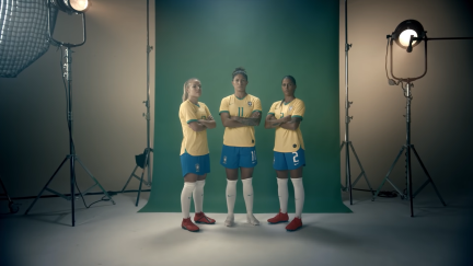 Captura de tela de um vídeo de propaganda. Três jogadoras de futebol posam de braços cruzados e expressão séria em um estúdio. Um fundo verde está posicionado atrás delas. Dois equipamentos de iluminação estão no lado direito e no lado esquerdo. A mulher mais à esquerda tem pele branca e cabelos loiros presos, as duas outras têm cabelos escuros presos. As três usam uniforme da seleção: camisetas amarelas, shorts azuis e meiões brancos. A do meio calça chuteiras brancas e as duas das pontas, vermelhas.