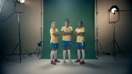 Captura de tela de um vídeo de propaganda. Três jogadoras de futebol posam de braços cruzados e expressão séria em um estúdio. Um fundo verde está posicionado atrás delas. Dois equipamentos de iluminação estão no lado direito e no lado esquerdo. A mulher mais à esquerda tem pele branca e cabelos loiros presos, as duas outras  têm cabelos escuros presos. As três usam uniforme da seleção: camisetas amarelas, shorts azuis e meiões brancos. A do meio calça chuteiras brancas e as duas das pontas, vermelhas.