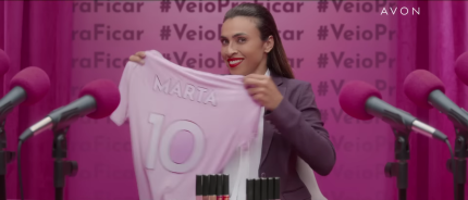Captura de tela de uma propaganda de batom com a jogadora de futebol Marta. A mulher tem pele negra, cabelo escuro e liso e olhos escuros. Ela usa camisa branca, blazer cinza e batom vermelho. Ela sorri enquanto segura e mostra uma camisa rosa com os escritos “Marta” e “10”. Batons estão posicionados à sua frente e nos cantos direito e esquerdo do primeiro plano estão microfones com espuma rosa. No fundo, cortinas cor de rosa com a hashtag “#VeioPraFicar”.