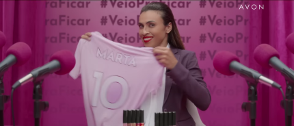 Captura de tela de uma propaganda de batom com a jogadora de futebol Marta. A mulher tem pele negra, cabelo escuro e liso e olhos escuros. Ela usa camisa branca, blazer cinza e batom vermelho. Ela sorri enquanto segura e mostra uma camisa rosa com os escritos “Marta” e “10”. Batons estão posicionados à sua frente e nos cantos direito e esquerdo do primeiro plano estão microfones com espuma rosa. No  fundo, cortinas cor de rosa com a hashtag “#VeioPraFicar”. 